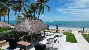 A beachside patio with thatched umbrellas, lounge chairs, and a view of the ocean. - Private Beach Retreat! Luxurious Bedding, Relaxing Pool, Best Sunsets, Fast WiFi (Key Colony Beach)