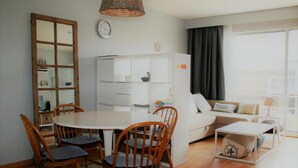 Una sala de estar moderna con comedor, muebles blancos, un ventanal grande y un reloj en la pared.