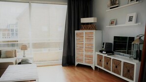 Una sala de estar con un ventanal grande, un armario blanco con cestas, un televisor y una lámpara.
