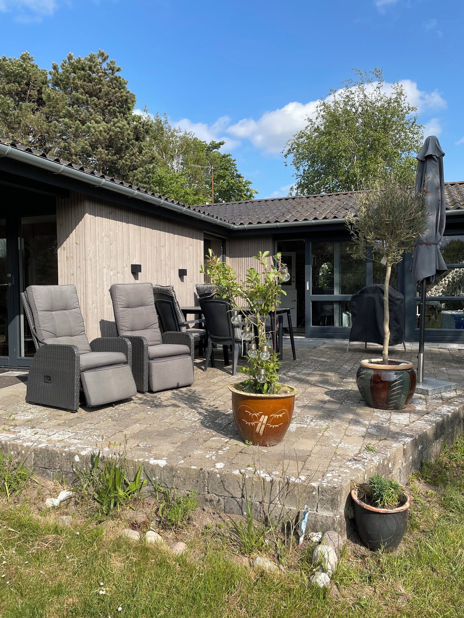 An outdoor seating area with cushioned chairs and tables, surrounded by potted plants and a building with a tiled roof.