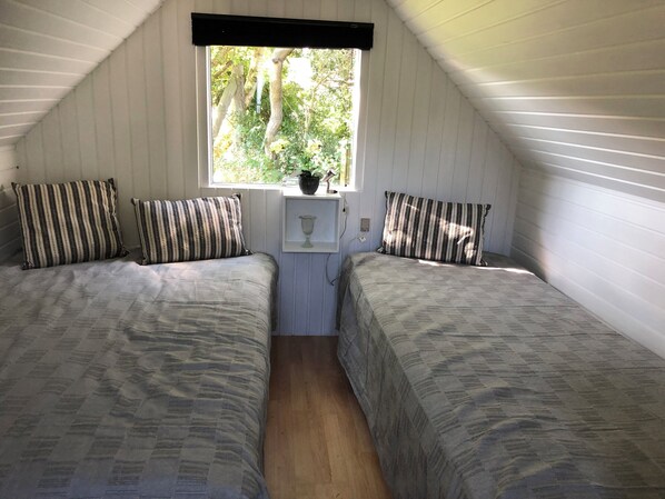 A small, cozy room with two beds, a window with a view of trees, and a small shelf with a vase.