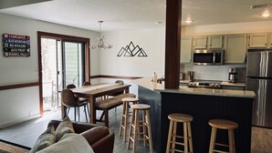 A modern kitchen with a central island, stainless steel appliances, and a dining area with a wooden table and bar stools. - Beautiful Camelback Views, Mountain - New Renovated 3-Bed / 2-bath home (Tannersville)