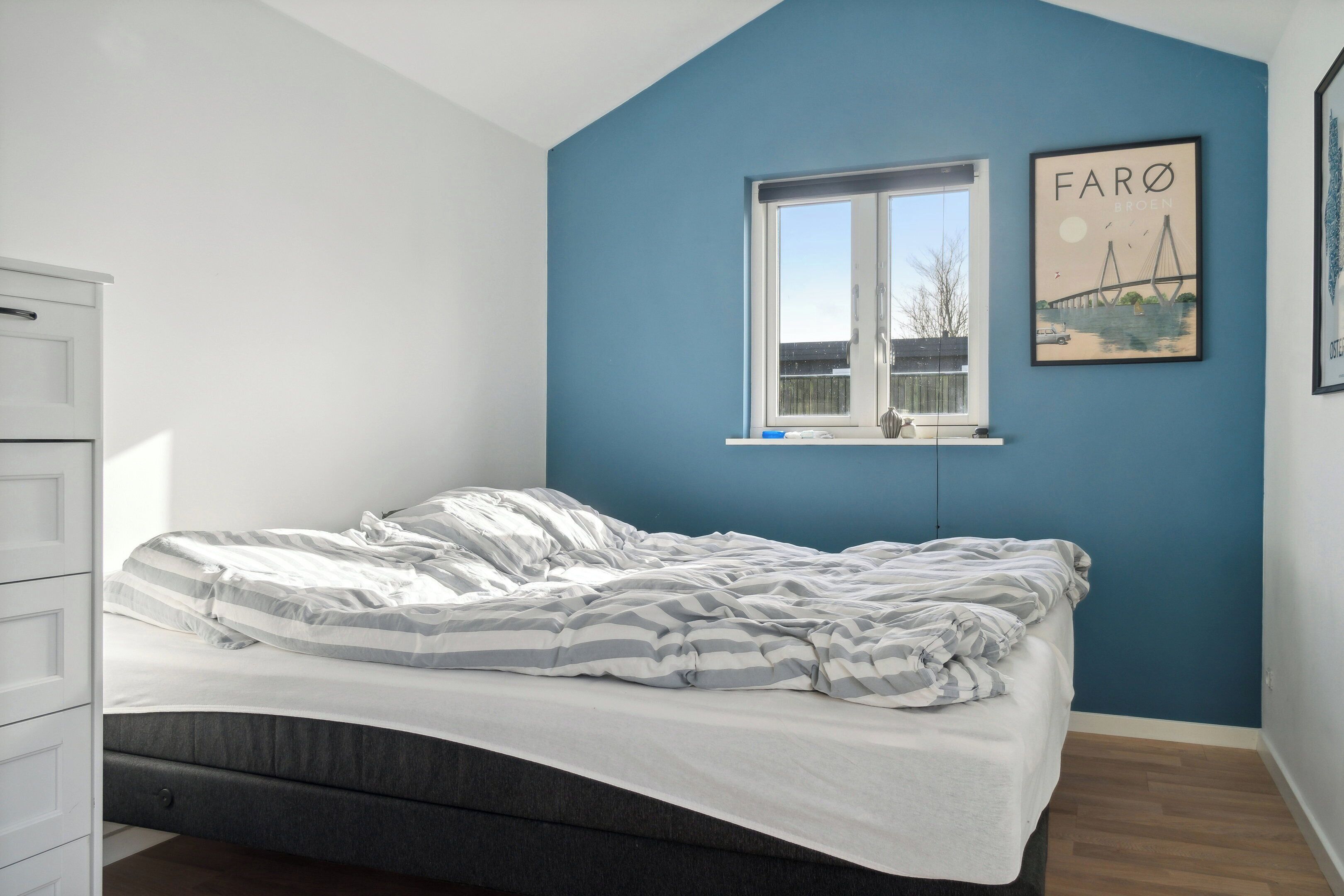 A bedroom with a bed, a window, a wall-mounted picture, and a white dresser.
