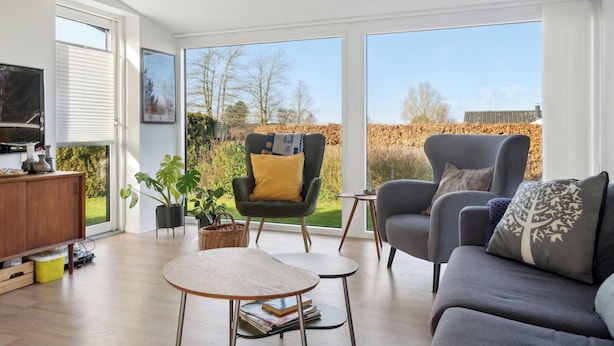 Una sala de estar moderna con sofá, sillón y mesa de centro, ventanas amplias y un televisor sobre un mueble de madera.