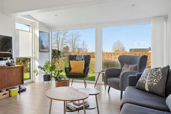 A modern living room with a sofa, armchair, and coffee table, large windows, and a television on a wooden cabinet.