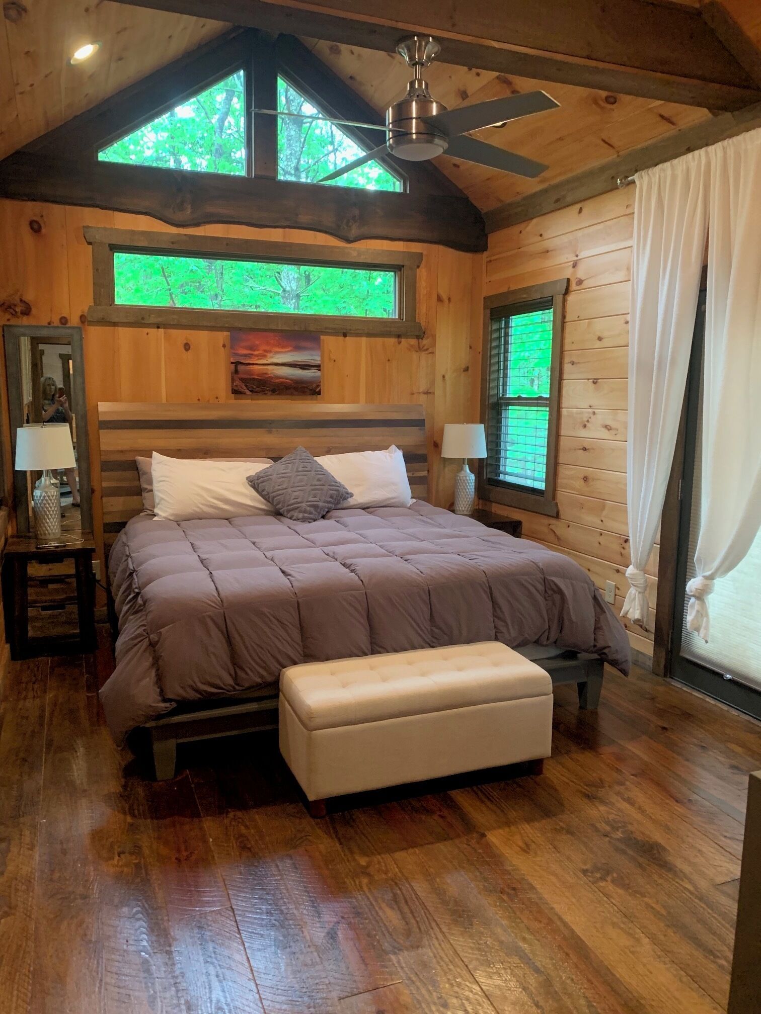 A wooden cabin bedroom with a bed, bench, ceiling fan, windows, and a painting.