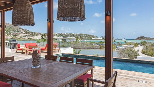 An outdoor dining area with a pool, wooden deck, wicker lamps, and a view of the ocean and hills.