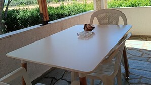 Un patio al aire libre con una mesa y sillas blancas, un adorno sobre la mesa, y una vista de vegetación y edificios al fondo.