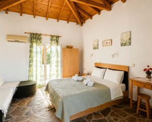 A bedroom with a stone floor, wooden ceiling beams, a bed with white linens, a wooden wardrobe, and a small table with a vase.