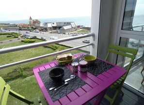 A balcony with a table set for dining, overlooking a coastal road and buildings.