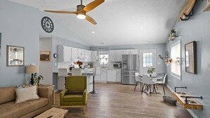 Une cuisine moderne avec des meubles blancs, un coin repas avec une table et des chaises blanches, et un salon avec un canapé et un fauteuil vert.
