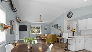 Une cuisine moderne avec des meubles blancs, un coin repas avec une table ronde et des chaises, et un salon avec un canapé et un fauteuil vert.