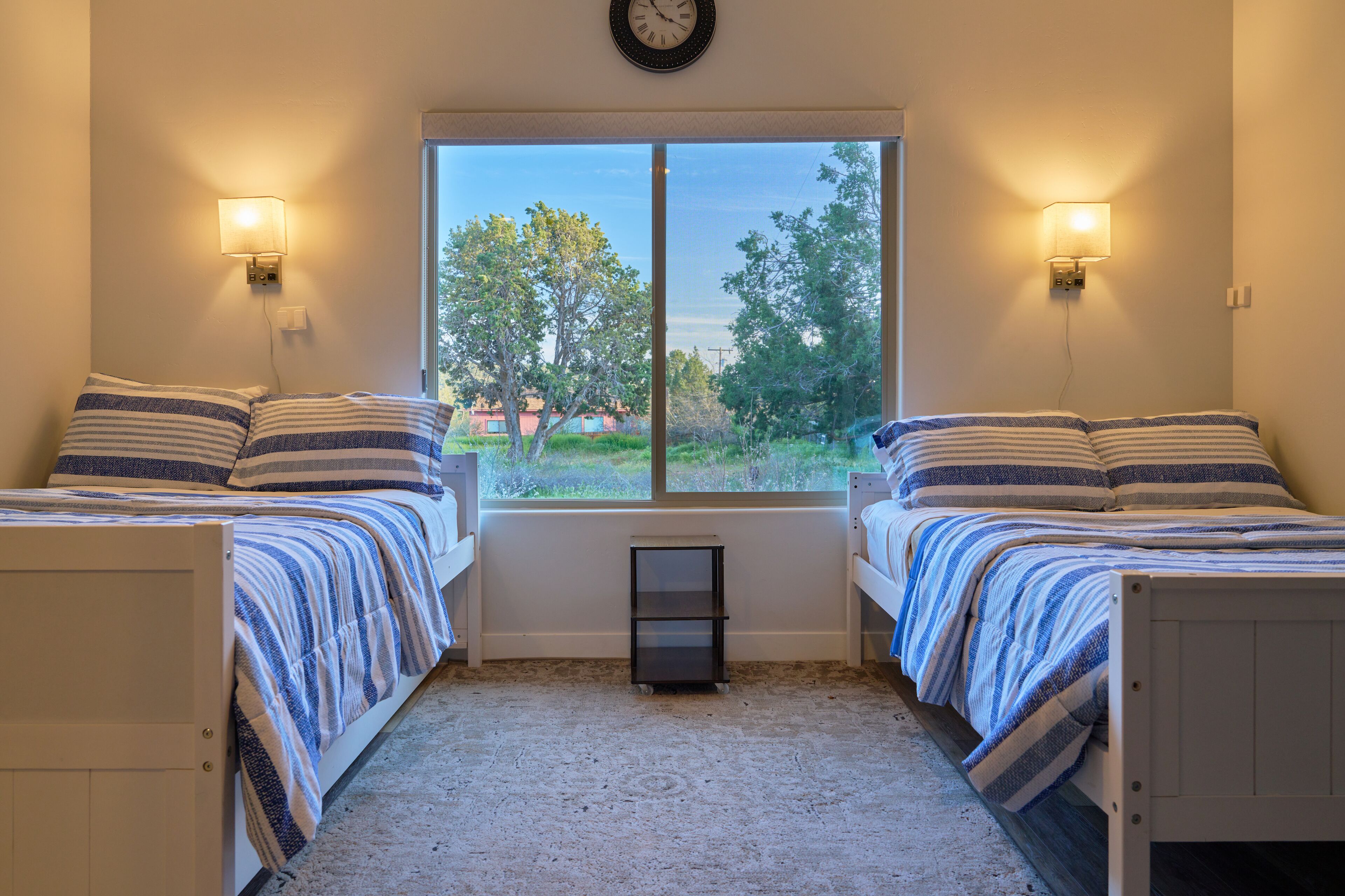 A bedroom with two beds, a window with a view of greenery, and wall-mounted lamps.