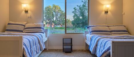A bedroom with two beds, a window with a view of greenery, and wall-mounted lamps.