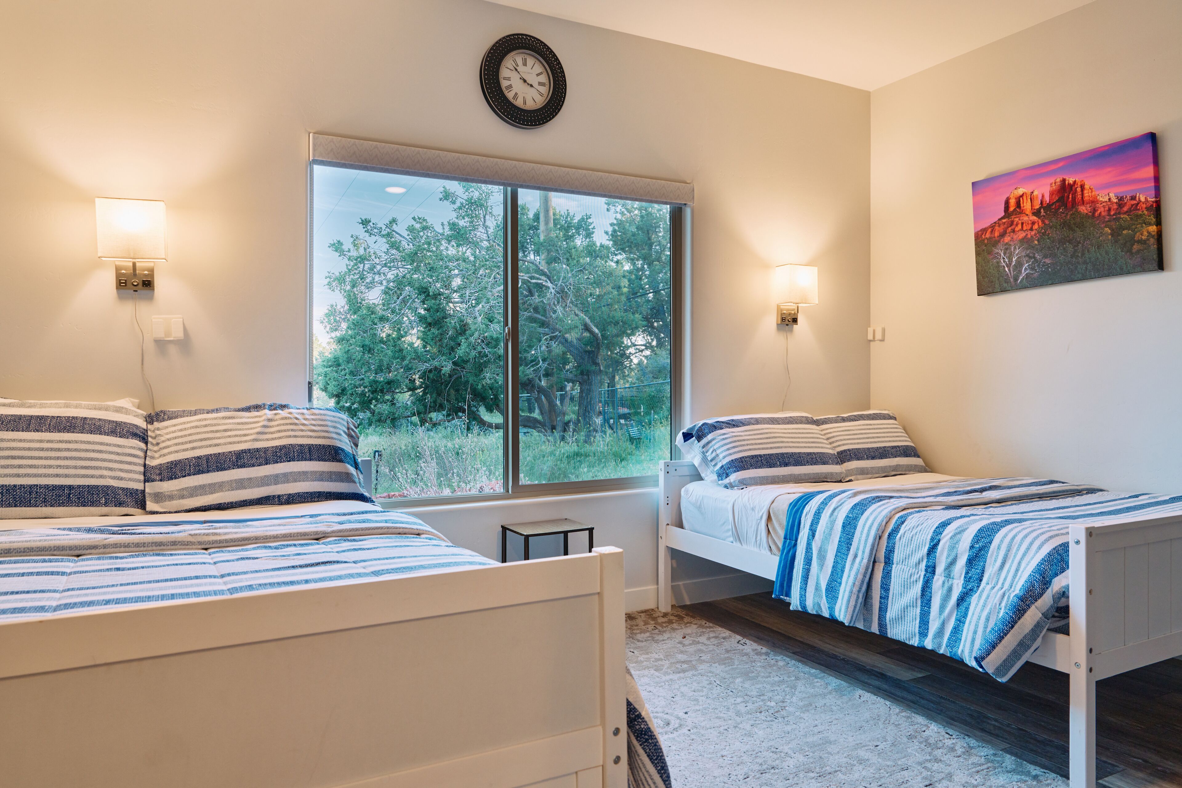 Un dormitorio con dos camas, una ventana con vista a árboles, un reloj y un cuadro en la pared.