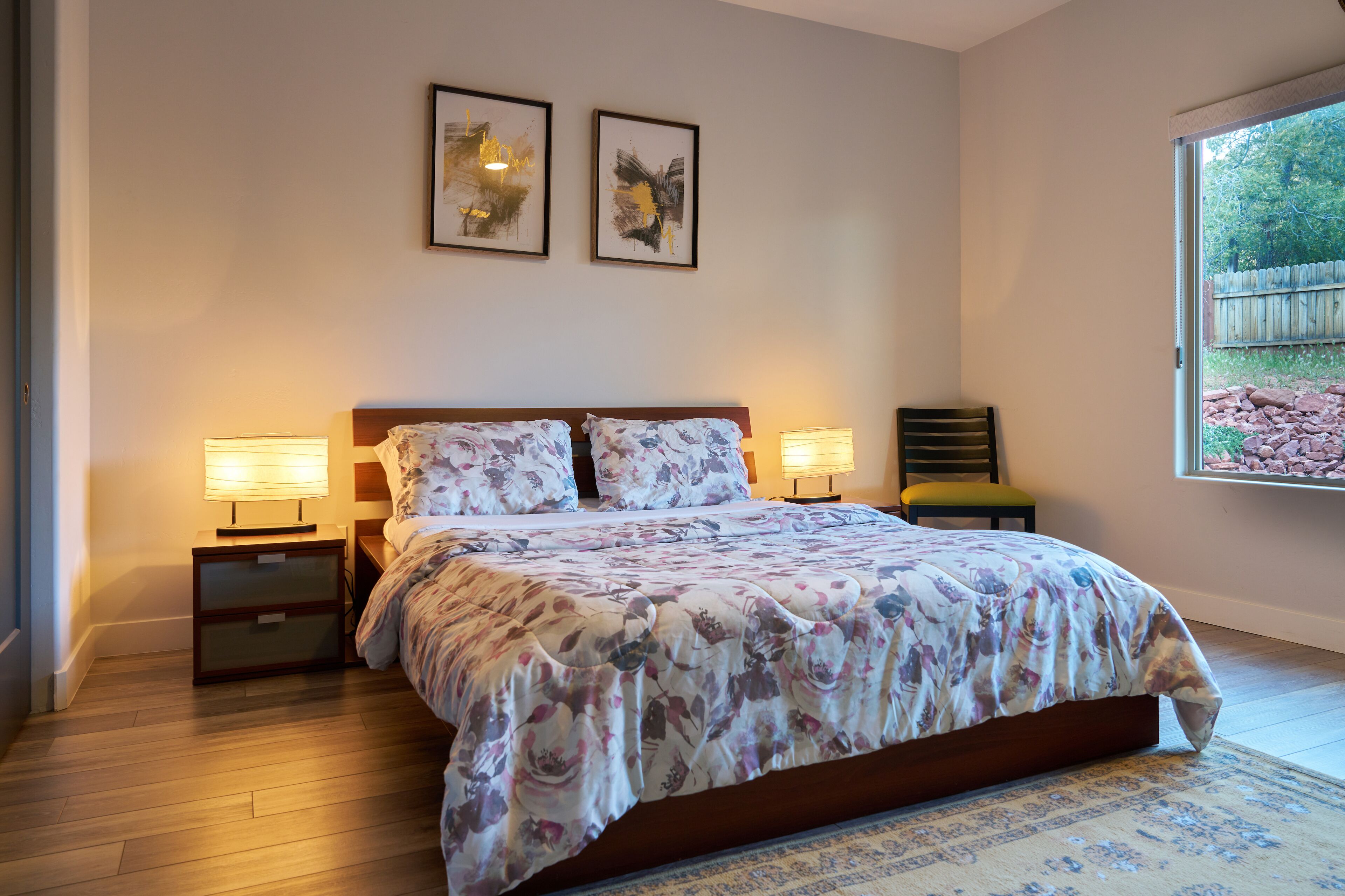 A bedroom with a bed, two bedside lamps, a nightstand, a chair, and a window with a view of trees.