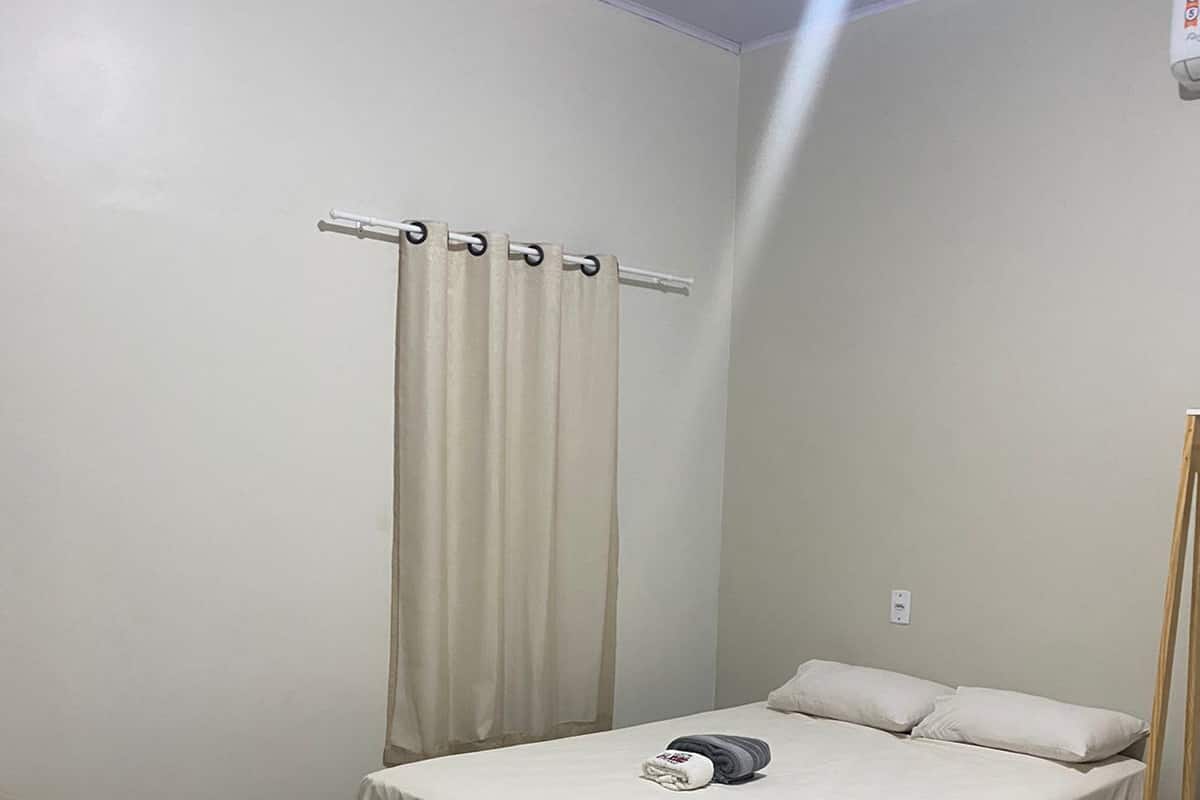A single bed with a white mattress and pillows, a small bedside table, and a curtain.