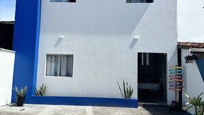 A two-story building with a white facade and blue trim. There are two windows on the ground floor and one on the upper floor. A sign with multiple destinations is visible in the entrance area. - Pousada e agência cledio turismo  (Barreirinhas)
