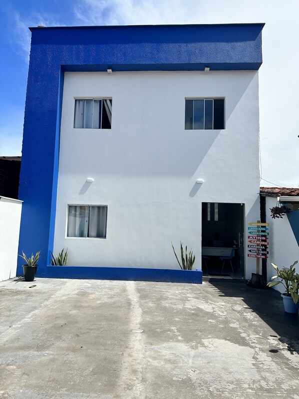 Ein zweistöckiges Gebäude mit weißer Fassade und blauen Verzierungen. Im Erdgeschoss befinden sich zwei Fenster, im Obergeschoss eines. Im Eingangsbereich ist eine Schilder mit verschiedenen Zielen zu sehen.