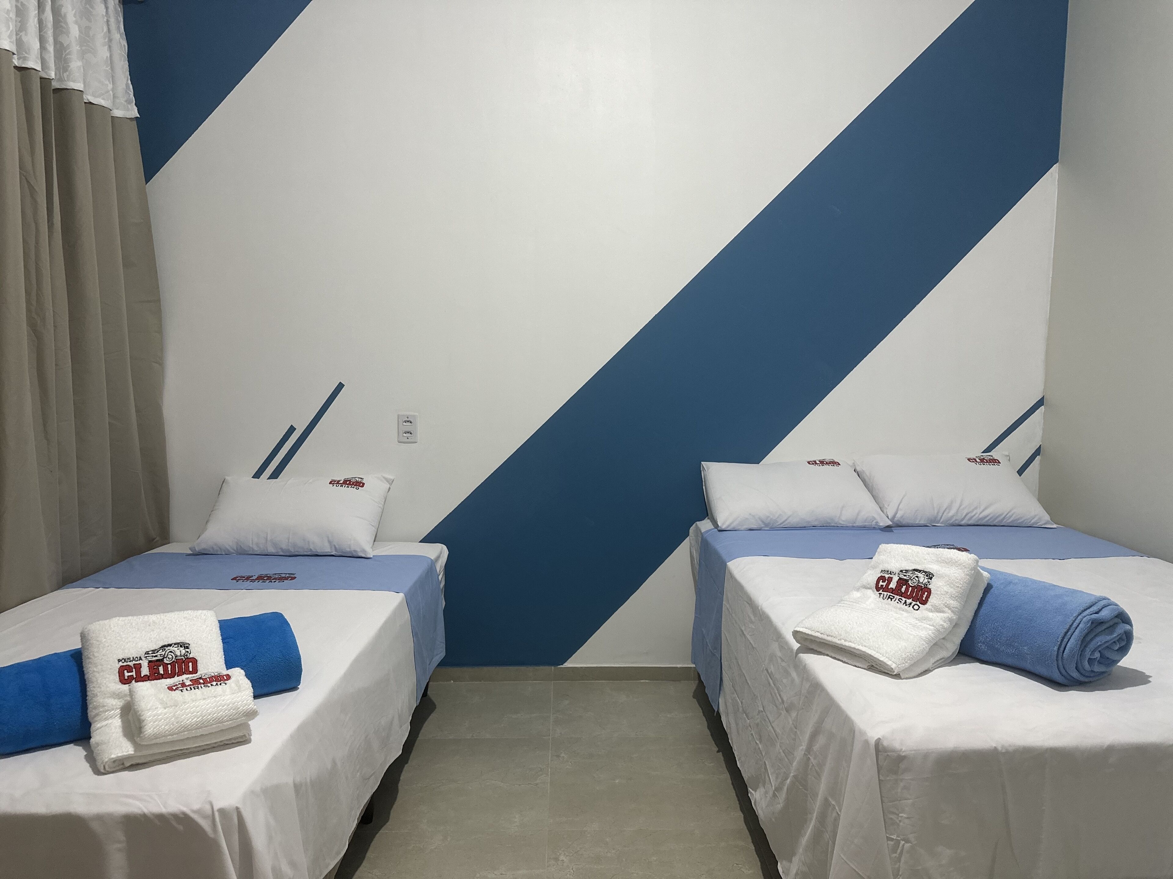 A hotel room with two single beds, each with a white bedspread, blue pillows, and a folded towel with text. The walls have a blue and white diagonal stripe pattern.