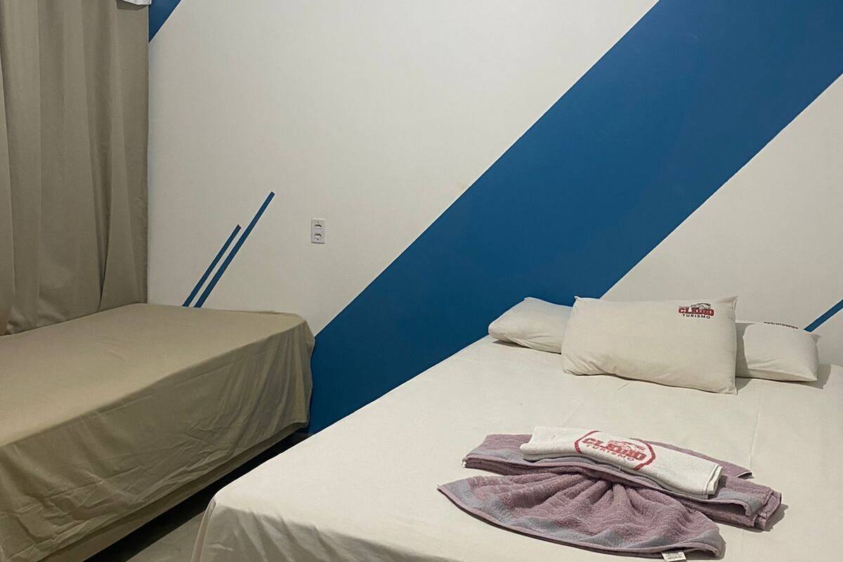 A room with two beds, one with a white bedspread and the other with a beige one. There is a blue and white wall design, a window with a white curtain, and a small table with a white tablecloth.