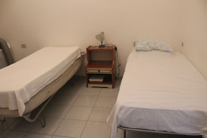 Two single beds with white linens, a wooden nightstand with a lamp, and a tiled floor.