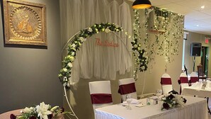 A banquet hall with round tables covered in white tablecloths, red and white chair sashes, and floral centerpieces. A decorative arch with greenery and lights is visible in the background.