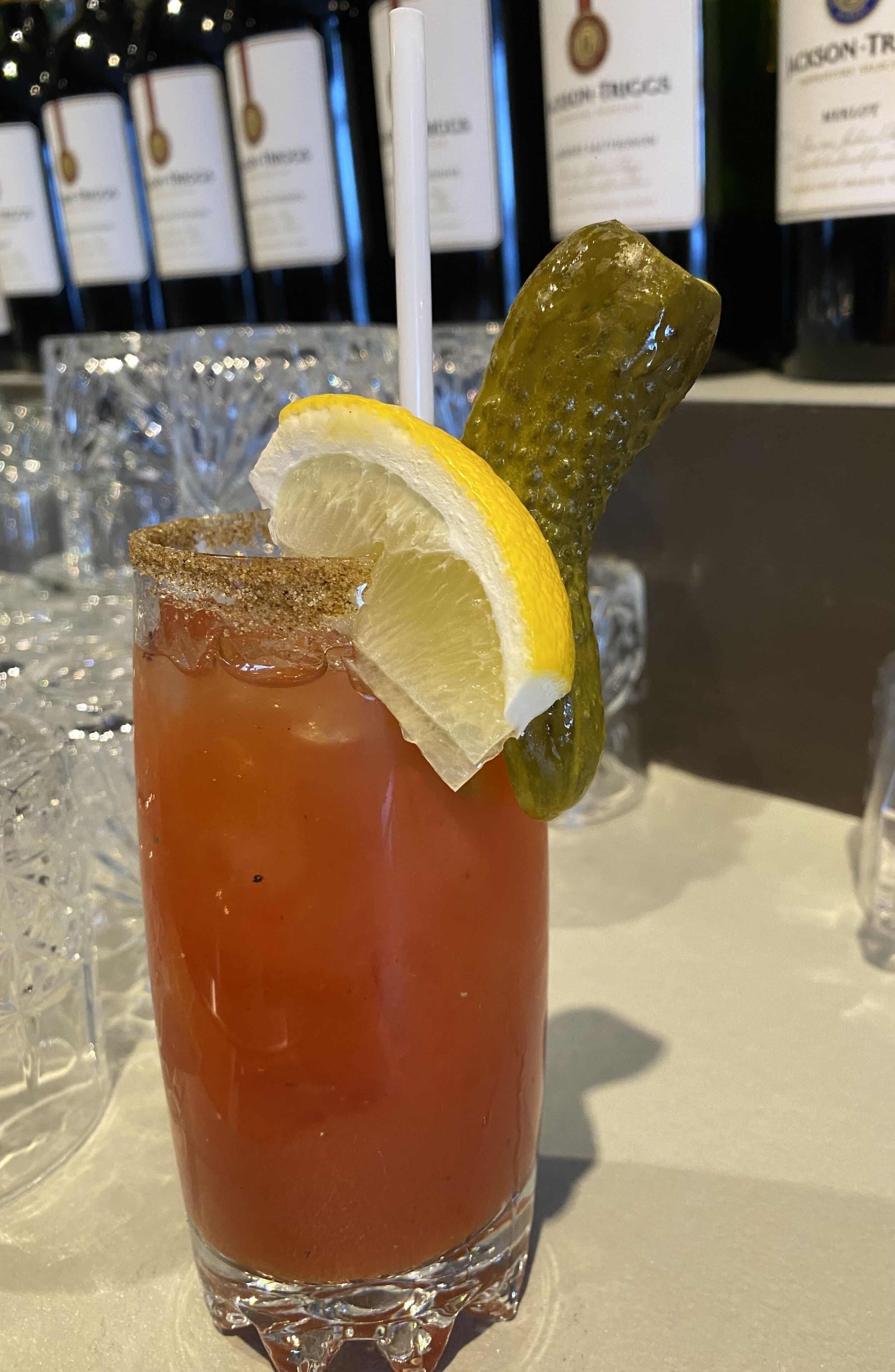 Bloody Mary met een citroenpartje en augurk, op een tafel met andere glazen en flessen op de achtergrond.