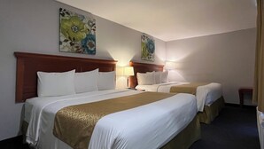 A hotel room with two beds, a wooden headboard, white bedding, a brown throw blanket, a nightstand with a lamp, and floral artwork on the walls.