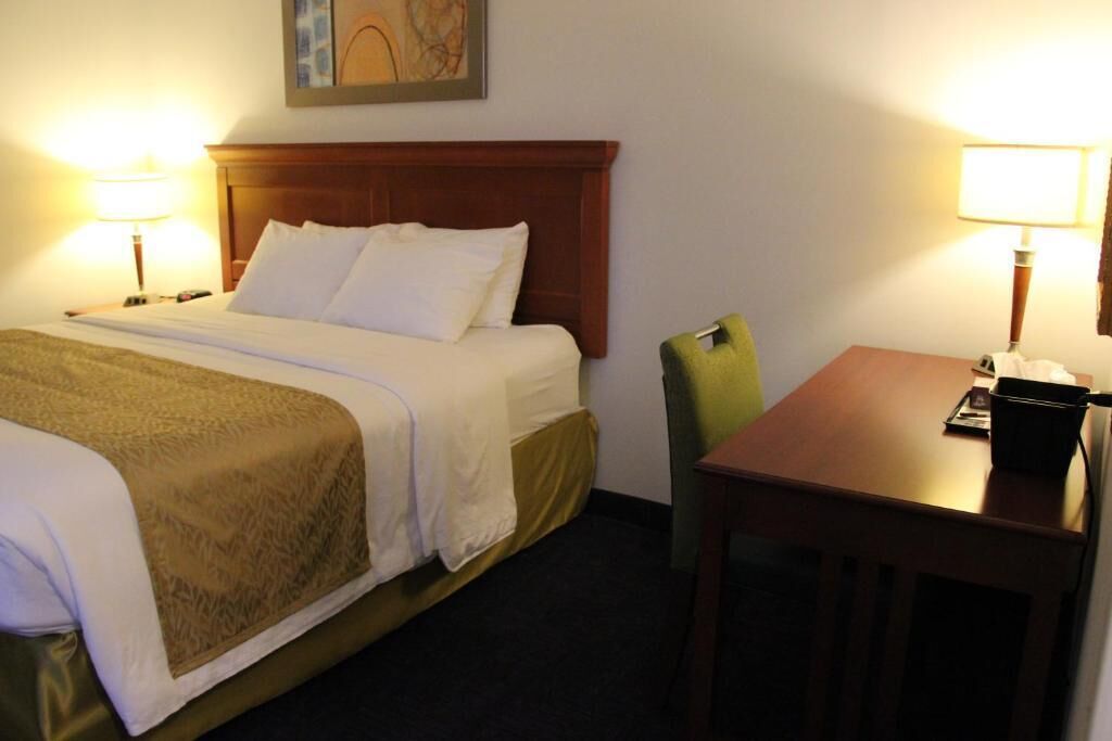 A hotel room with a bed, two bedside lamps, a wooden headboard, a green chair, and a small table with a lamp.