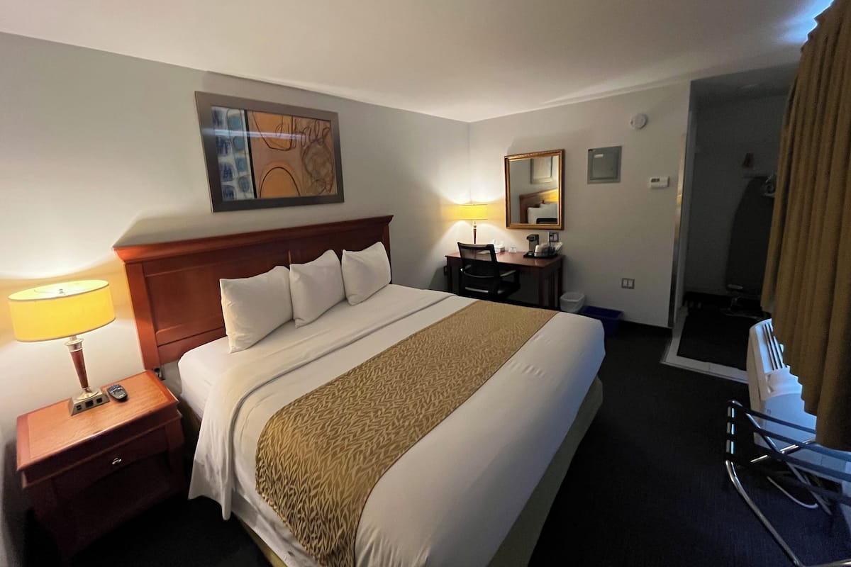 Habitación de hotel con una cama grande, un escritorio con silla, una lámpara, un espejo y zona de ducha.