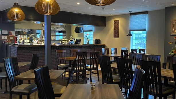A restaurant interior with wooden tables and chairs, hanging pendant lights, a bar area, and a view of the street through large windows.