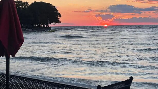 Ein Sonnenuntergangsblick von einer Terrasse am Meer mit Tisch und Stühlen, einem roten Sonnenschirm und einer entfernten Küstenlinie.