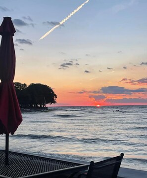 A sunset view from a seaside terrace with a table and chairs, a red umbrella, and a distant shoreline. - Angola on the Lake- Grandview Bay-25 minutes from Highmark Stadium (Angola)