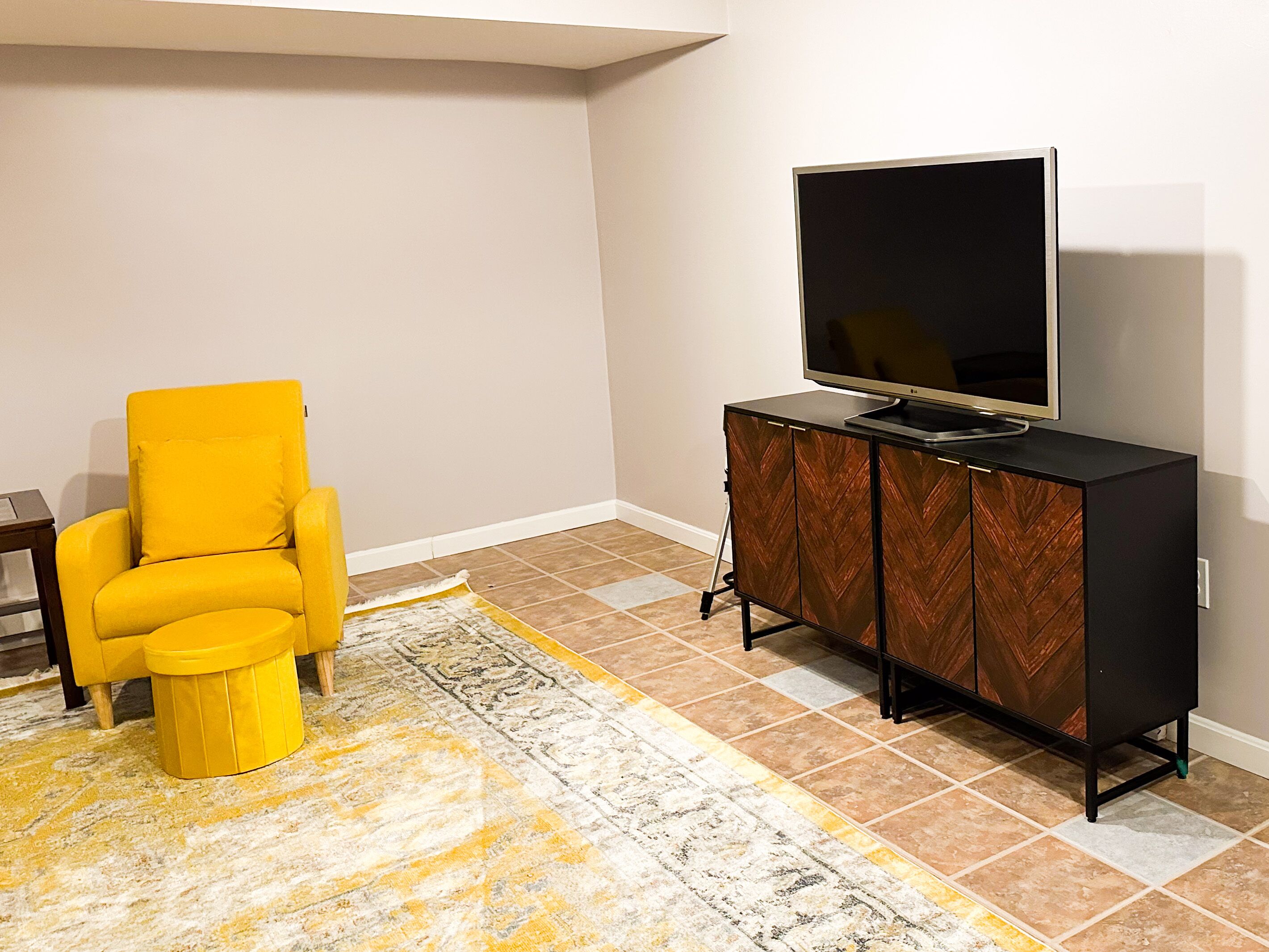 Un salon avec un fauteuil jaune, un téléviseur à écran plat posé sur un meuble noir et un tapis à motifs.