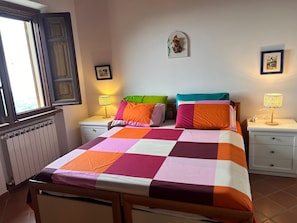A bedroom with a checkered bedspread, two bedside tables with lamps, a window with a wooden frame, and a wall-mounted picture.