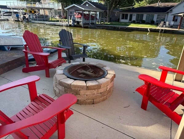 A fire pit surrounded by red Adirondack chairs on a concrete deck by a body of water.