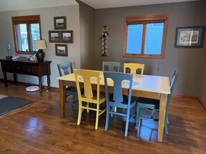 A dining area with a wooden table and chairs, framed pictures on the wall, and a window with blinds.