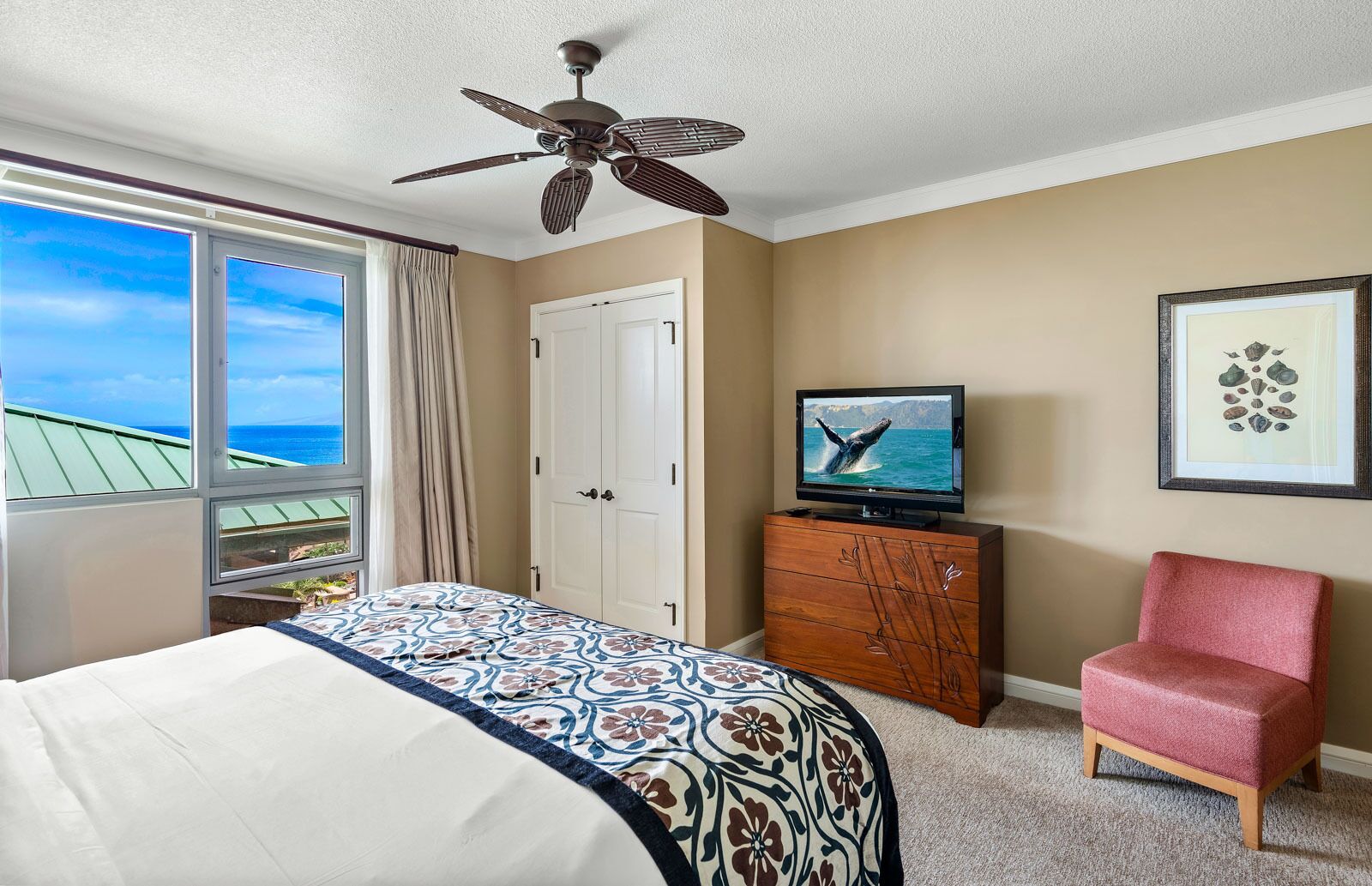 A hotel room with a bed, a television, a chair, a ceiling fan, and a window with a view of the ocean.