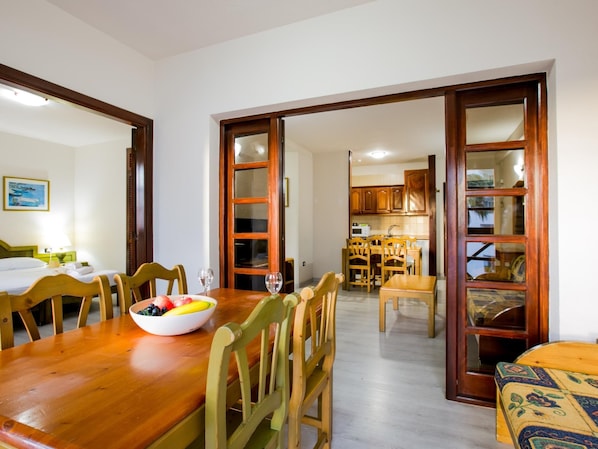 Un coin repas avec une table en bois, des chaises et un saladier rempli de fruits. On peut apercevoir une cuisine à travers une porte ouverte.