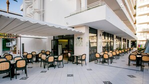 An outdoor cafe area with tables and chairs, a covered seating area, and a building with multiple floors and balconies. - 104 Andorra close to the beach (Playa de las Américas)