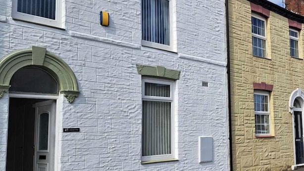 Una hilera de casas urbanas de dos pisos con fachadas blancas y amarillas, una puerta visible y una pequeña ventana con un objeto amarillo en la pared.