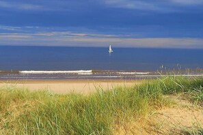 A beach with a sailboat in the distance, grassy dunes in the foreground, and a clear sky. - Cosy 2-Bed Cottage - Free WiFi (Bridlington)
