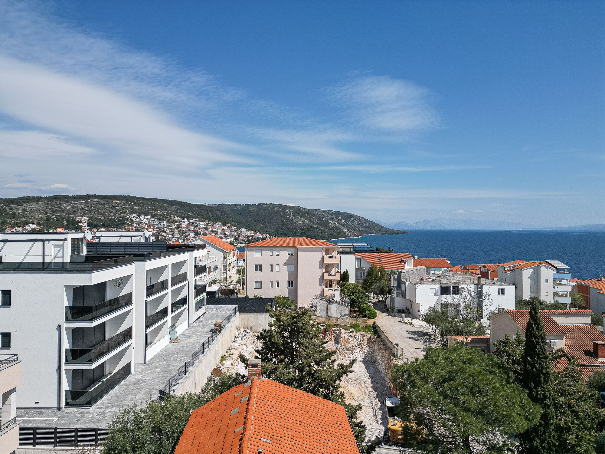 A coastal town with modern buildings, a clear view of the sea, and a hilly landscape in the background.