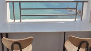 A balcony with two wooden chairs and a white table, overlooking a clear blue sea and distant land.