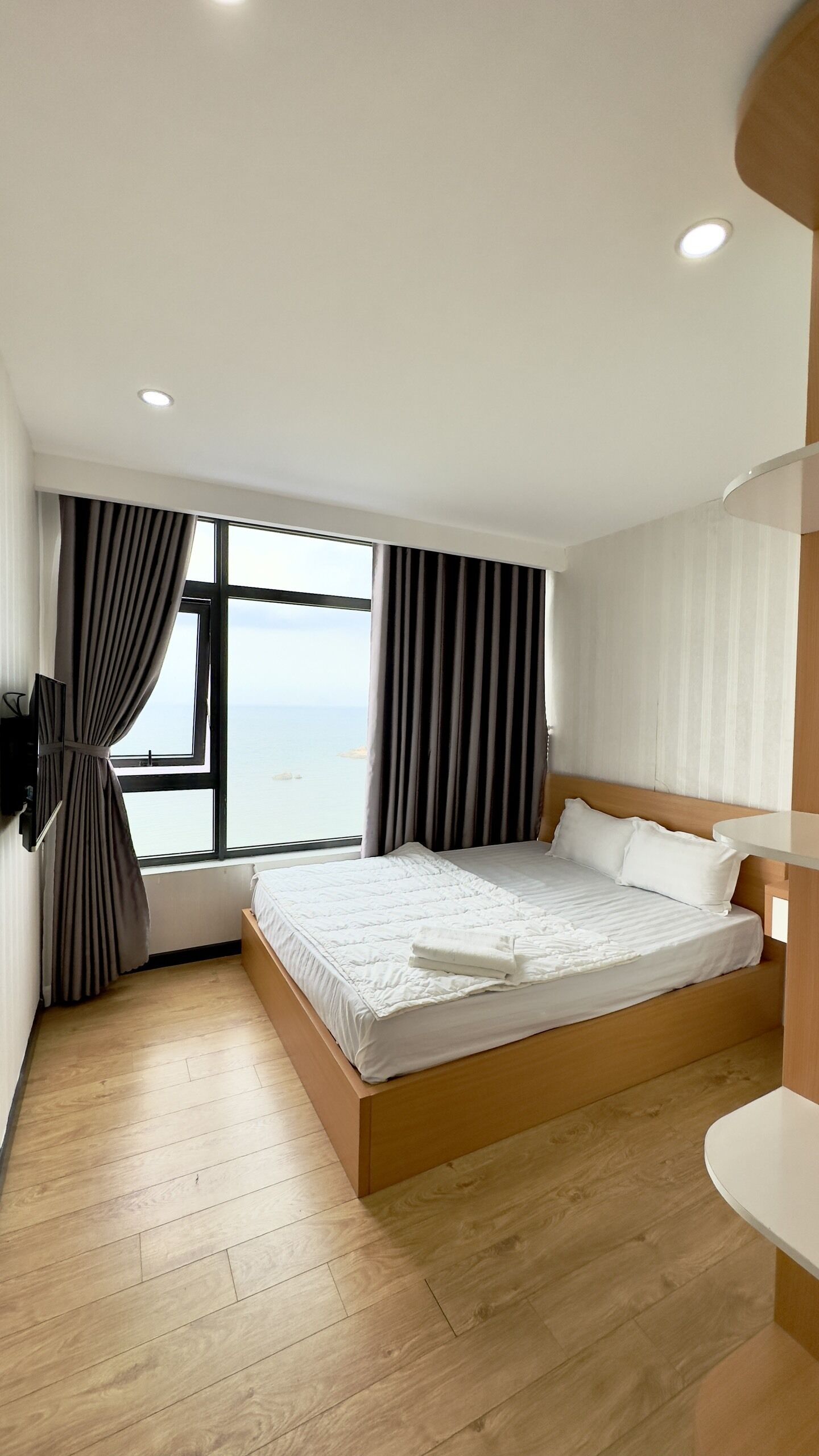 A modern hotel room with a large bed, wooden flooring, a view of the sea, and a flat-screen TV mounted on the wall.