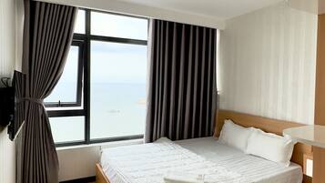 A modern hotel room with a large bed, wooden flooring, a view of the sea, and a flat-screen TV mounted on the wall.