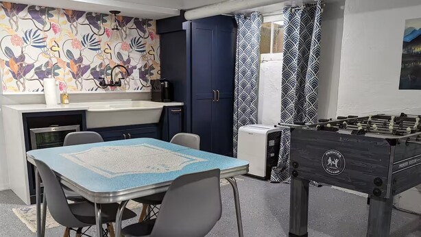 Um quarto com mesa de pebolim, mesa de jantar com cadeiras, área de cozinha com pia e armários, e uma parede com padrão.