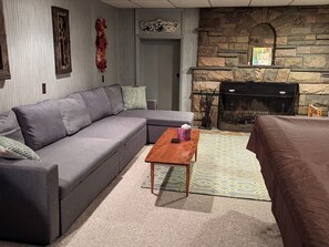 A living room with a grey sofa, a wooden coffee table, a patterned rug, a stone fireplace, and a door leading to another room.
