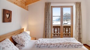 Un dormitorio con techo de madera, una cama con ropa de cama floral, una mesita de noche, una decoración con forma de corazón y una ventana con vistas a un paisaje nevado.